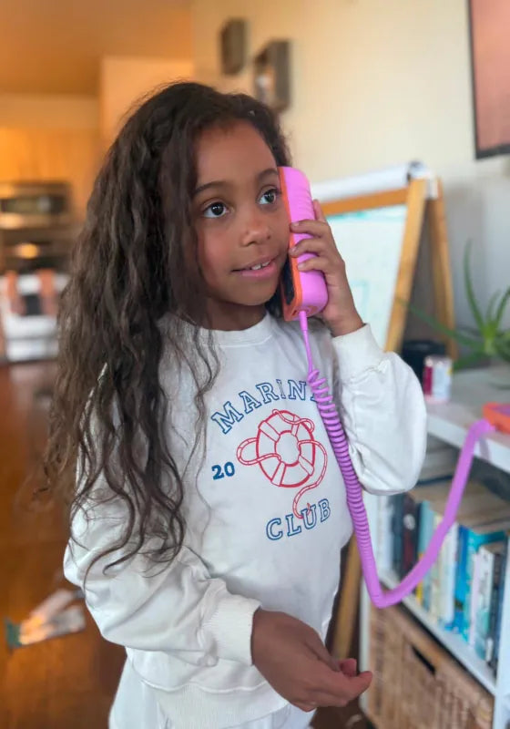A girl in a Marine Club sweatshirt talking on a Later Alligator Lilac Tin Can phone for kids