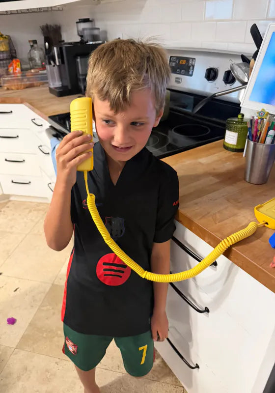 A boy in the kitchen holding a Landline Lemon Tin Can handset with its twirly cord