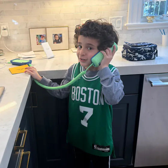 A boy in a Boston jersey talking on an Answer Me Aquamarine Tin Can in the kitchen