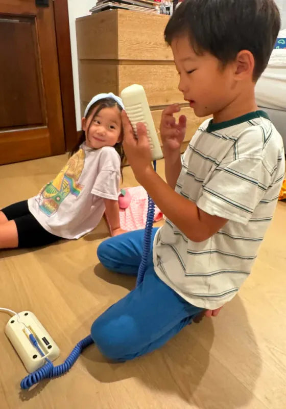 A boy holding a Static White Tin Can handset for a younger girl as they sit on the floor