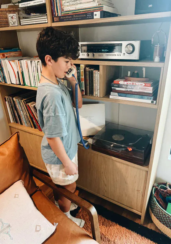 A boy by a bookshelf and record player, talking on a Static White Tin Can with its twirly cord